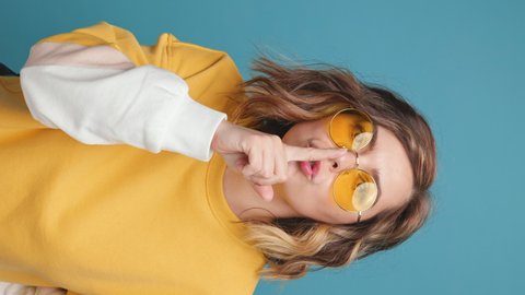 Vertical video Silence gesture Happy Young Woman showing brings index finger to lips makes gesture for silence sunglasses, smiles looks at camera on blue background. Don't tell expression. Lifestyle