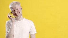 Positive Albino Guy Talking With Banana Cellphone Having Fun Posing Standing Over Yellow Studio Background. Mobile Communication, Phone Call Concept. Advertisement Banner With Empty Space For Text
