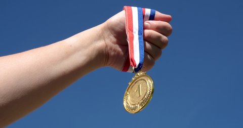 A hand holds up a first-place gold medal, with the sky in the background. Victory concept