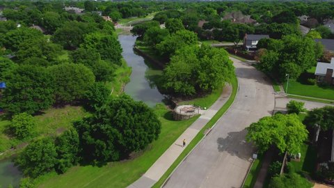 An aerial shot of a Dallas neighborhood park with a person walking on a path next to a river as the drone flies forward following the street as a car drives by