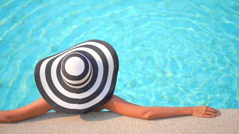 Back of a woman in Floppy stripped hat leaning on pool border inside swimming pool, template copy space