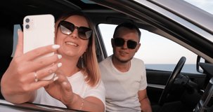 Young Man and Woman Sitting in Car with Sea View from Window and Using Smartphone for Video Call, Waving Hands to Say Hello During Their Road Trip