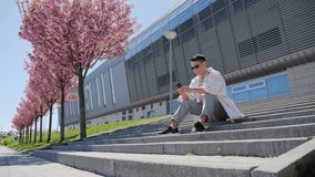  Asian Attractive man in Casual Clothes and Sunglasses Sitting on the Stairs near Big Modern Office Building with Beautiful Trees Having some Rest Chatting Messaging using App of Smartphone outdoors.
