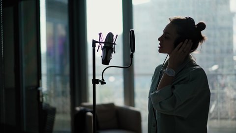 A young woman with headphones sings into a microphone on a stand with a pop filter, singer records a song at a recording studio.