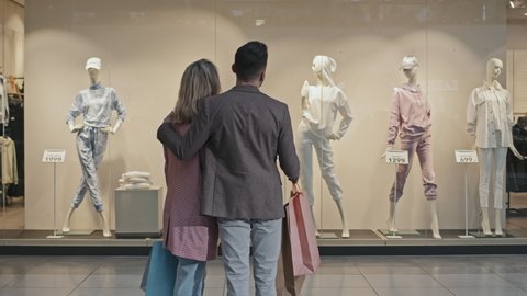 Medium stab rear-view shot of young stylish couple shopping for clothes together walking along big modern shopping mall standing in front of window display to look at mannequin
