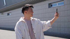 Young confident Asian Man in Casual Clothes Having Video Call with Friend near Modern Building and flowering trees Using his Smartphone. Social Network. Work outside. Success. People. Travel.