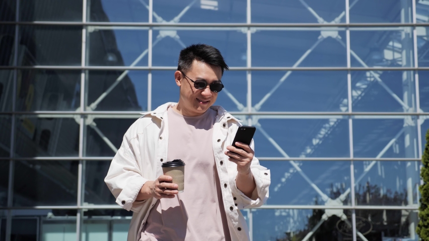 Successful happy Asian Businessman in Casual Clothes is texting Messages on modern Smartphone. Young Man is walking in Business Quartal, having good Mood, looking Satisfied. Successful Lifestyle. Men. Royalty-Free Stock Footage #1072792532