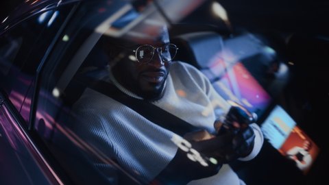 Stylish Black Man in Glasses is Commuting Home in a Backseat of a Taxi at Night. Handsome Male Using Smartphone and Looking Out of Window while in a Car in Urban City Street with Working Neon Signs.
