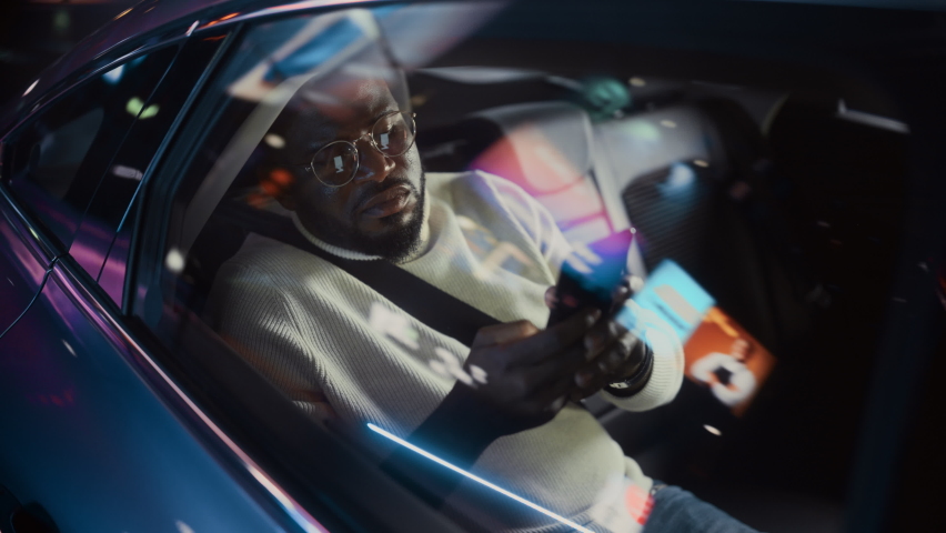 Stylish Black Man in Glasses is Commuting Home in a Backseat of a Taxi at Night. Handsome Male Using Smartphone and Looking Out of Window while in a Car in Urban City Street with Working Neon Signs. - Powered by Shutterstock - Get 15% off with code: PIKWIZARD15