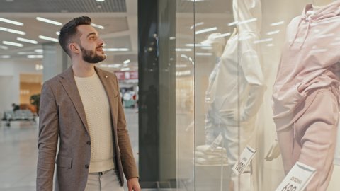 Medium shot of handsome bearded man in smart clothes carrying lots of shopping bags walking along shopping mall looking at mannequins in window displays