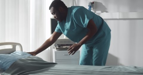 African male nurse changing sheets in hospital ward. Portrait of afro-american medical staff making bed and cleaning in clinic. Healthcare and hygiene concept