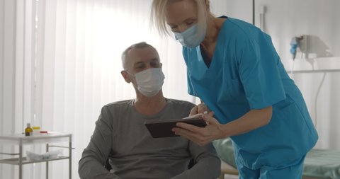 Nurse wearing medical mask giving documents to aged patient in wheelchair to sign. Senior disabled man in wheelchair wearing protective mask and signing consent in hospital ward
