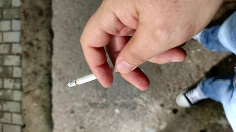 Close-up vertical video of a man's hand holding a smoking cigarette. Man's hand holding a burning cigarette while smoking, tobacco smoke addiction, unhealthy lifestyle concept.