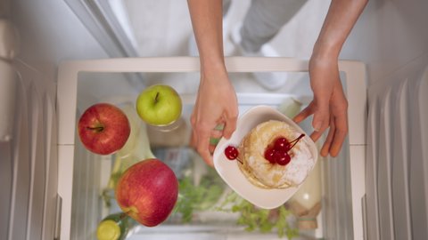 Woman's hands choosing between apples and cake from refrigerator. Top View from inside the working fridge.
