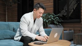 Chinese businessman sitting on the couch typing on laptop keyboard while having video call with business partners in his cozy loft office. Smiling asian man having online video conference in office.