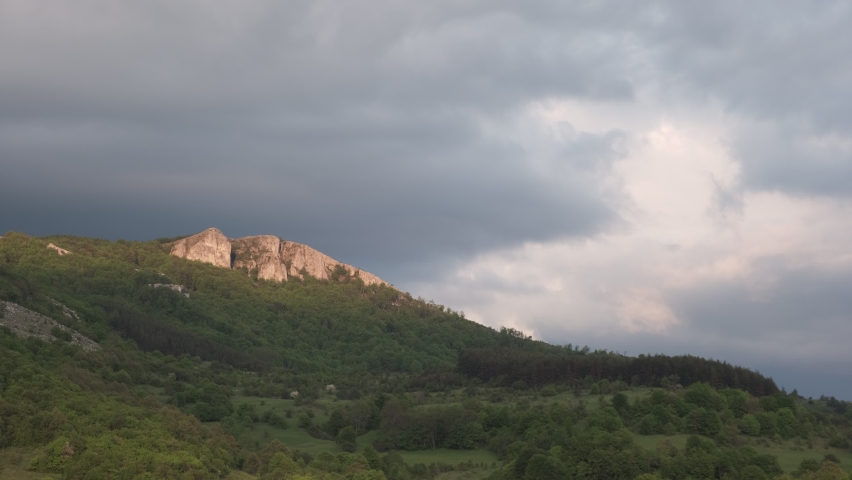 Alpenglow on a pointy, rocky peak during sunset on a mountain covered by vivid green fields and forests during early spring and a cloudy sky
