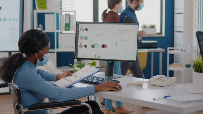 Black woman with disabilities and protection face masks working on computer in new normal workplace during pandemic. Multiethnic team in business financial office checking reports, analysing datas - Powered by Shutterstock - Get 15% off with code: PIKWIZARD15