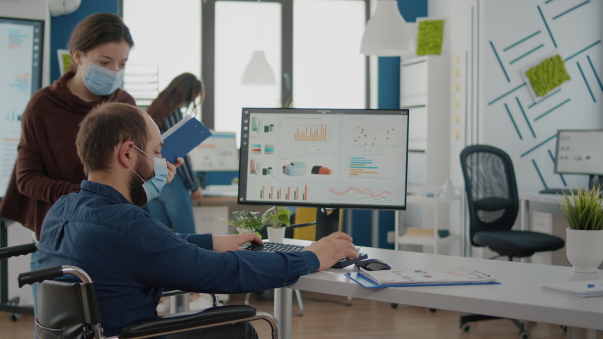 Invalid worker in wheelchair talking with colleague, working start up office wearing face masks during covid 19 pandemic. Team in new normal business financial company typing on pc, checking reports - Powered by Shutterstock - Get 15% off with code: PIKWIZARD15