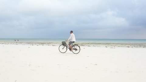 4K footage of a man dressed light summer clothes riding old vintage bicycle with front basket on the lonely low tide ocean white sand coast on Kiwengwa beach on Zanzibar island, Tanzania.