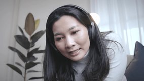 Happy cheerful Asian woman wearing wireless headphones recording video for social media sitting on the sofa at home lifestyle and pleasure