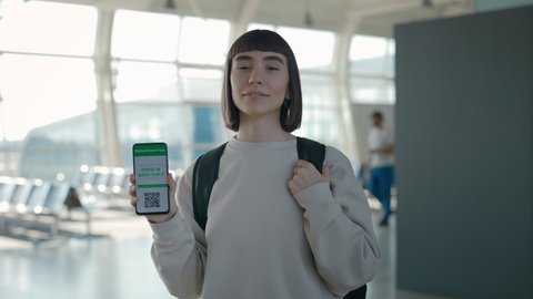 Portrait of charming lady holding modern smartphone with certificate with immunity for Covid-19 on screen. Female tourist with backpack using digital green pass while travelling abroad.