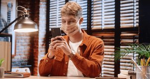 Young stylish guy student in protective face mask sits in cozy apartment spending free time playing online games on his smartphone.