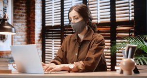 Beautiful stylish woman sitting alone at home in self-isolation in protective mask and working on laptop remotely.