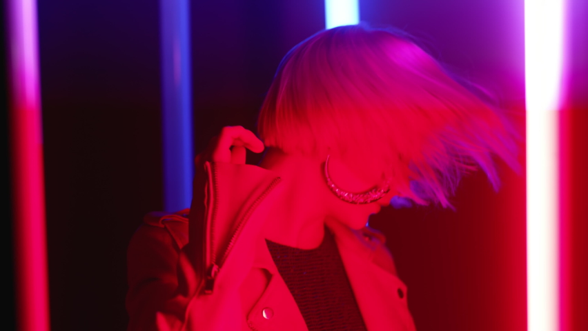 Fashionable woman with dyed pink hairstyle dancing relaxed in room with colorful neon glowing lamps. Hipster with trendy make-up. Night club, futuristic outfit. Sexy lady concept. 