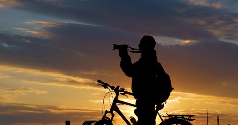 Silhouette of male photographer cyclist stands with camera on road and looks into the distance and admires the beautiful landscape at crimson sunset.  