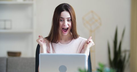 Excited young woman looking at laptop screen, celebrating win, showing yes gesture, sitting at desk in office home, rejoicing success, reading good news email, online lottery victory, actively dance