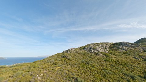 View from above, flying at high speed over a mountain range covered by a green vegetation with the mediterranean sea in the distance. Sardinia, Italy. Drone point of view, mountain surfing.