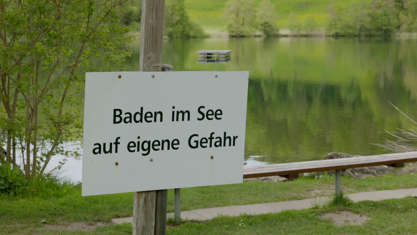 lake outdoor pool with swimming platform in green landscape with notice sign in german, german text translation: swim in the lake at your own risk at the lake, camera tilt to rescue swim ring