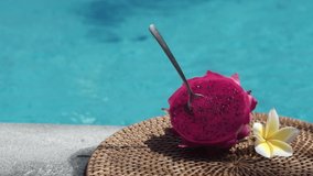 Fresh red dragon fruit cut into two halves with a spoon stuck into the pulp on the edge of bubbling blue swimming pool with white tropical flowers frangipani, moving shade from palm branch in Bali