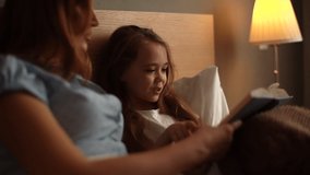 Close-up portrait of happy young mother and adorable daughter reading together children book before going to sleep while lying in bed in dark nursery bedroom, near lamp. Tracking shot in slow motion. - Powered by Shutterstock - Get 15% off with code: PIKWIZARD15