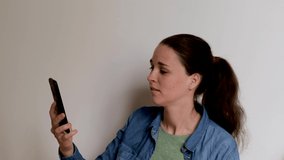 Young Caucasian European woman on white background is talking on video link using smartphone and looks at phone screen carefully. 4K footage. Conversation over Internet with business partner.