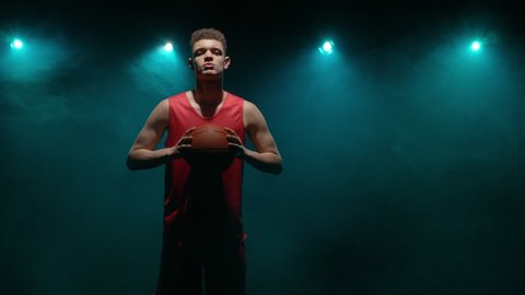 Out of darkness and smoke, silhouette of guy walking straight into camera with basket ball in his hands looms. Basketball player posing in studio with back blue light. Slow motion ready 59.94fps.