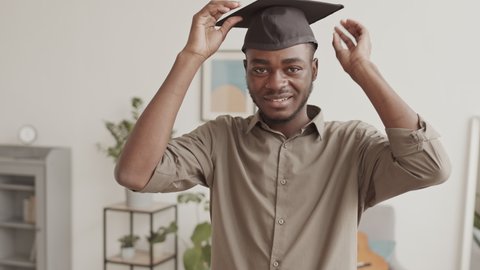 Medium shot of young African-American man standing in living room, putting on graduation hat while looking at camera, pointing at camera and saying joyful words