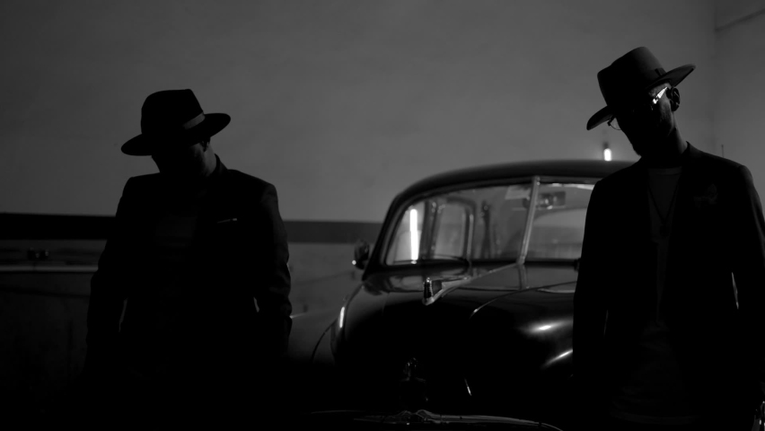 Two silhouettes of men in black suits with classic hats standing by vintage old car. Strangers look like italian mafioso from movie . Footage have permissible count of normally film effect style GRAIN Royalty-Free Stock Footage #1072887515