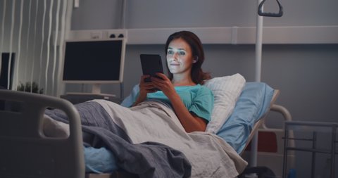 Beautiful young patient woman lying in hospital bed and using digital tablet. Portrait of sick woman resting in clinic ward surfing internet on tablet computer