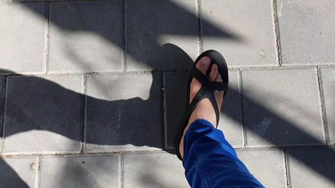 Female legs in blue pants and feet in black sandals while walking straight ahead on sidewalk, shot from above. Closeup of woman's lower body part pacing fast. Shadow on left side.