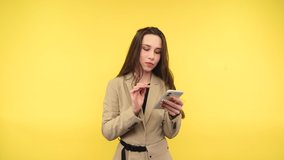 Beautiful woman in smart clothes uses a smartphone on a yellow background, looks at the screen with a calm face, wears a beige jacket.