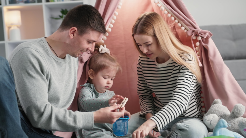 family fun, joyful woman and man playing and developing motor skills of cute little girl with wooden clothespins sitting on background of wigwam at home