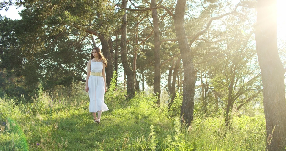 A cheerful teenage girl in light dress walks in a clearing between green trees in summer forest.
