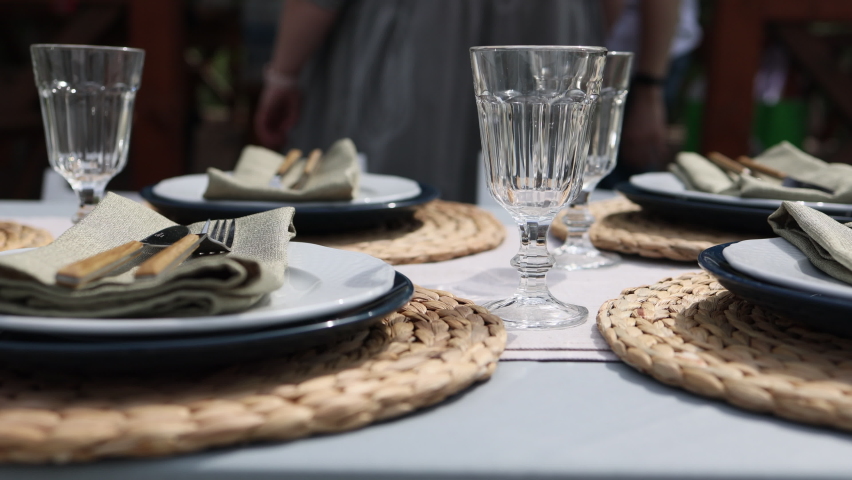 Laconic, minimalistic table setting. White plates on knitted napkins, glass glasses. Dinner table for eight people in nature. Sunny day. Celebrated. | Shutterstock HD Video #1072901954