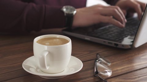 The man is drinking coffee. Work at the computer and drink a fresh cup of coffee. A man works at a laptop while drinking coffee from a white cup. Solid wood textured table close-up