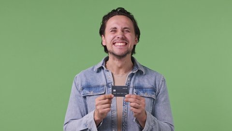 Handsome european friendly happy young man 20s years old wears denim shirt pointing index finger on mockup plastic credit bank card showing thumbs up isolated on pastel green color background studio