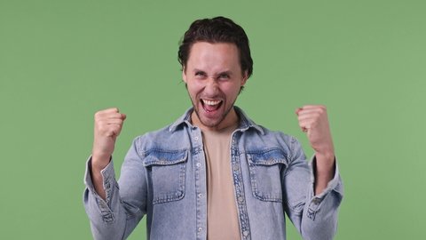 Excited fun jubilant overjoyed happy brunet blue eyed young man 20s years old wears denim shirt doing winner gesture celebrate clenching fists say yes isolated on pastel green color background studio
