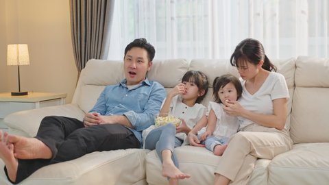 Happy Asian family stay home, Father and Mother spend leisure time with little two young daughters in living room. Parents and small preschool girl sit on sofa, watching movie on TV together in house.