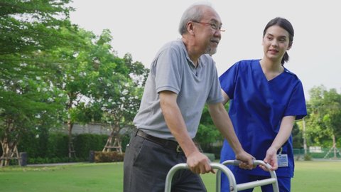 Asian young woman nurse at nursing home take care disabled senior elderly man. Caregiver doctor serve physical therapy for older patient to exercise and practice walking on walker or cane at backyard.