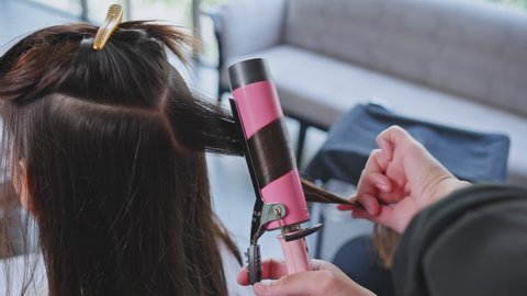 Close up of Expert man stylist's hand use curling iron make curly to young girl's hair in beauty salon. Unrecognize woman customer looking at mirror enjoy new hairstyle from hairdresser at barber shop